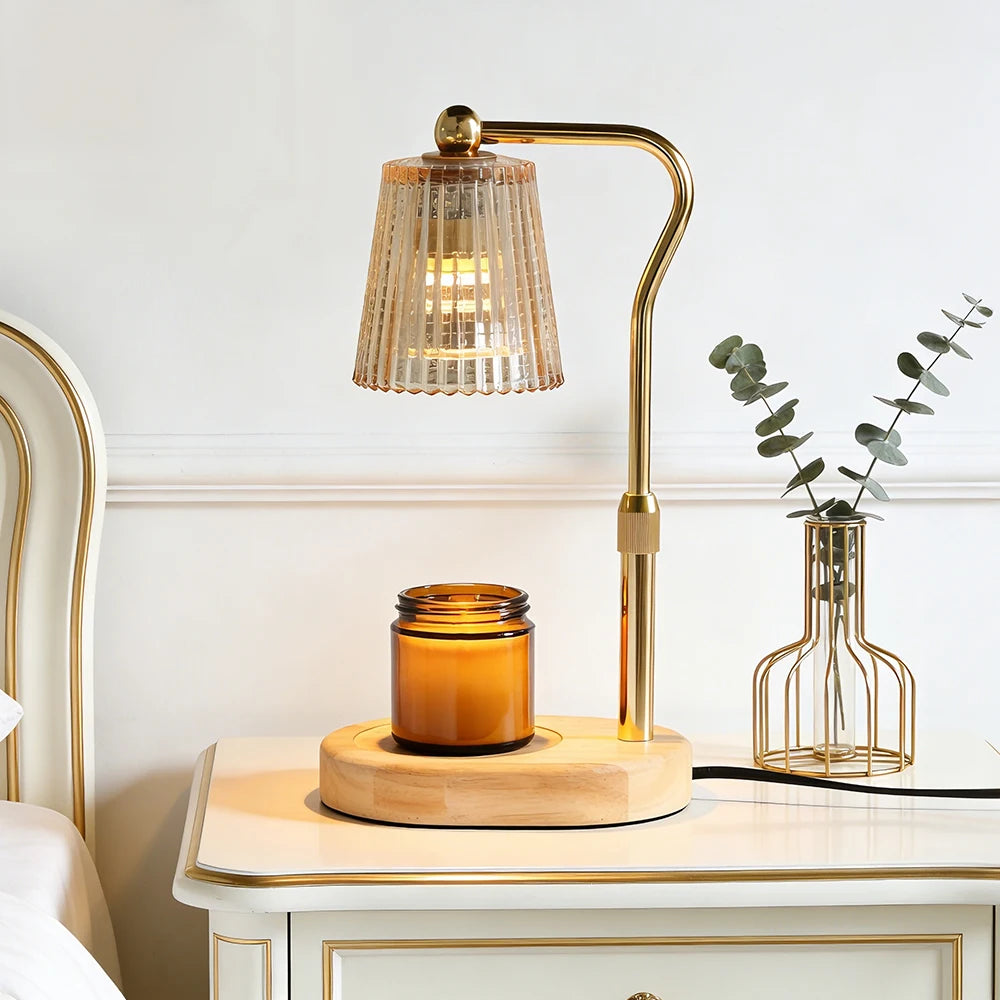 A modern table lamp with a curved brass stand and a ribbed clear glass shade, shown lit on a white nightstand beside a small vase with eucalyptus. The lamp base sits on a round wooden tray next to an amber glass jar candle.