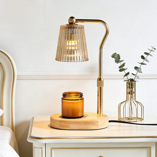 A modern table lamp with a curved brass stand and a ribbed clear glass shade, shown lit on a white nightstand beside a small vase with eucalyptus. The lamp base sits on a round wooden tray next to an amber glass jar candle.