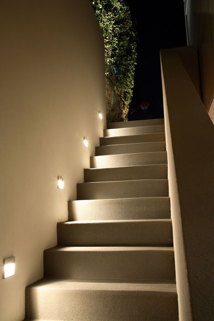 Outdoor staircase with warm under-stair lighting, beige stone steps, a beige wall with recessed lights, and a green hedge at the top against a dark night sky.