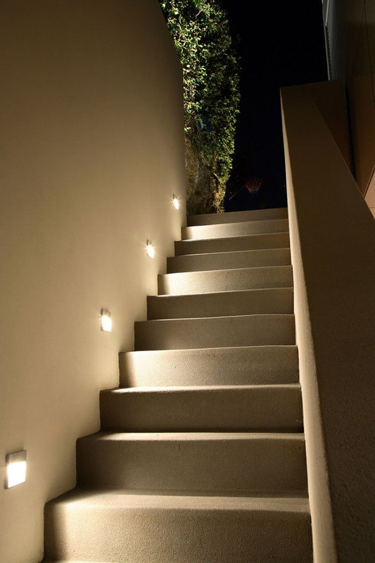 Outdoor staircase with warm under-stair lighting, beige stone steps, a beige wall with recessed lights, and a green hedge at the top against a dark night sky.